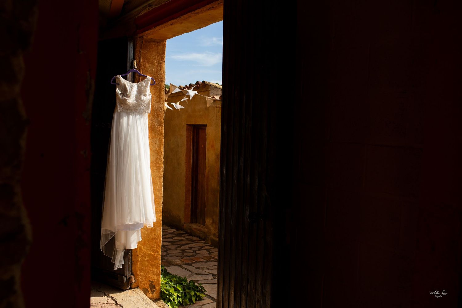 Elegant white wedding dress hangs in a rustic doorway with warm adobe walls and blue sky.