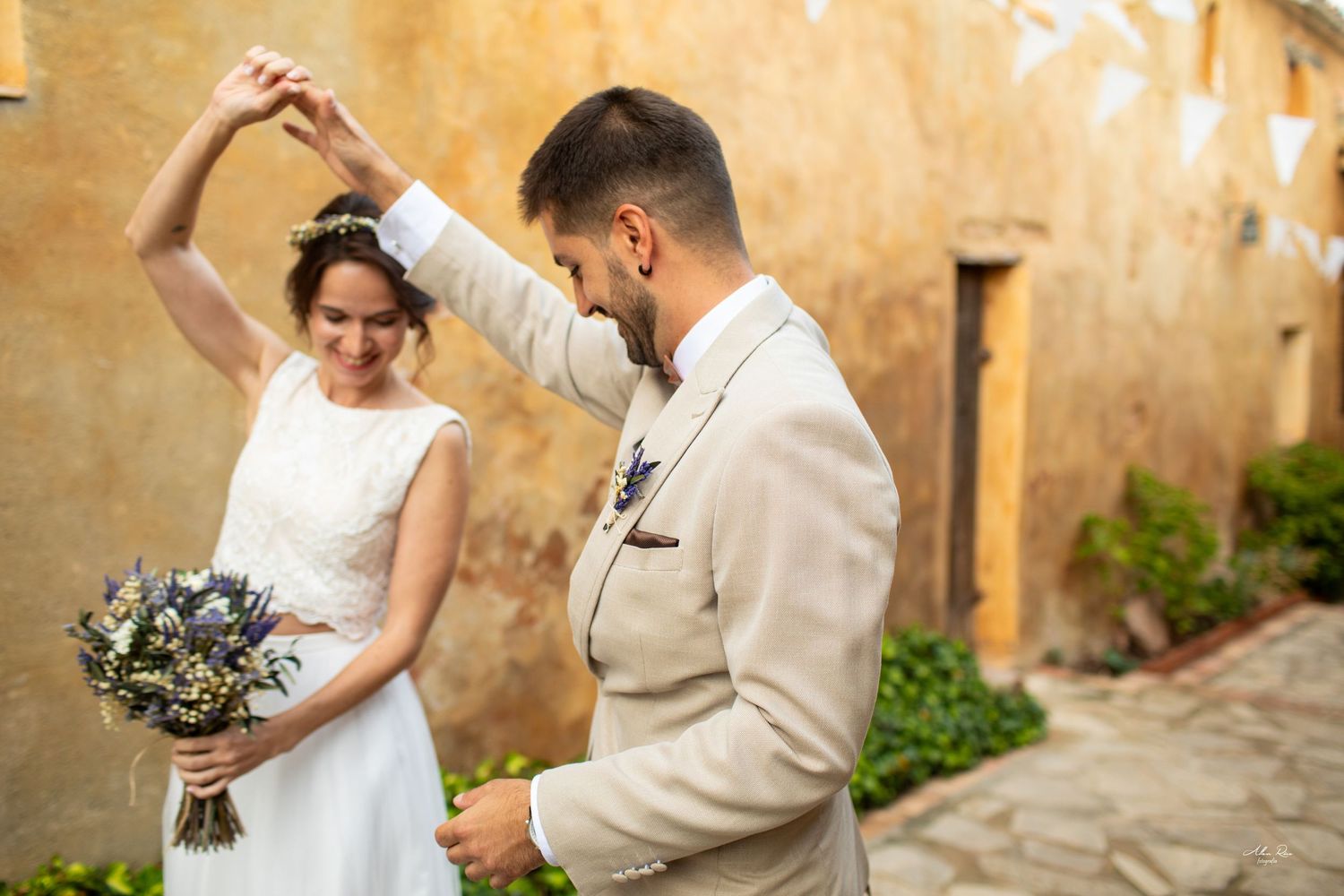Groom spins bride holding wildflower bouquet in joyful moment against golden textured wall.