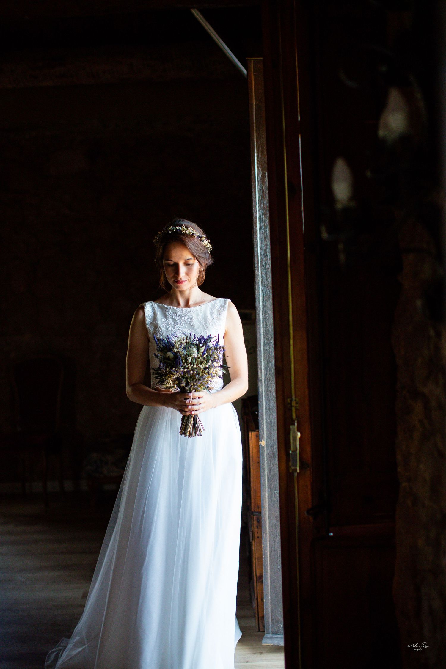 Elegant bride in white tulle gown with floral crown holds bouquet in rustic doorway.