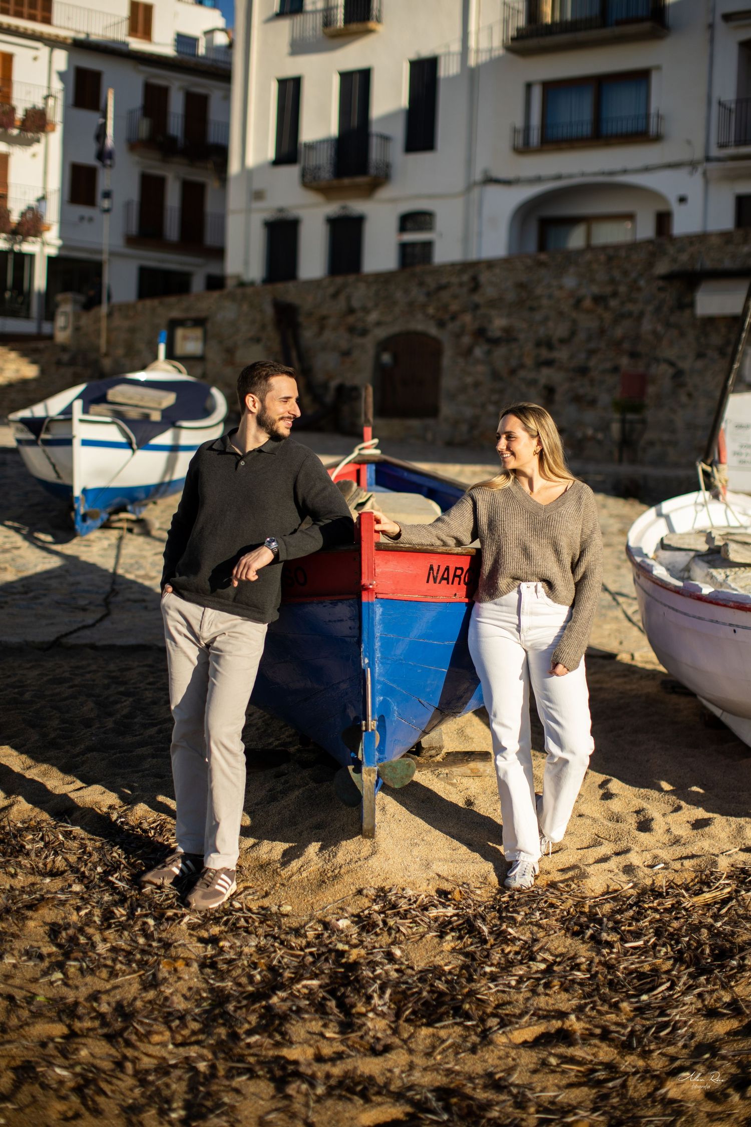 Couple smiling at each other beside a colorful red and blue boat on a sandy Mediterranean village beach.