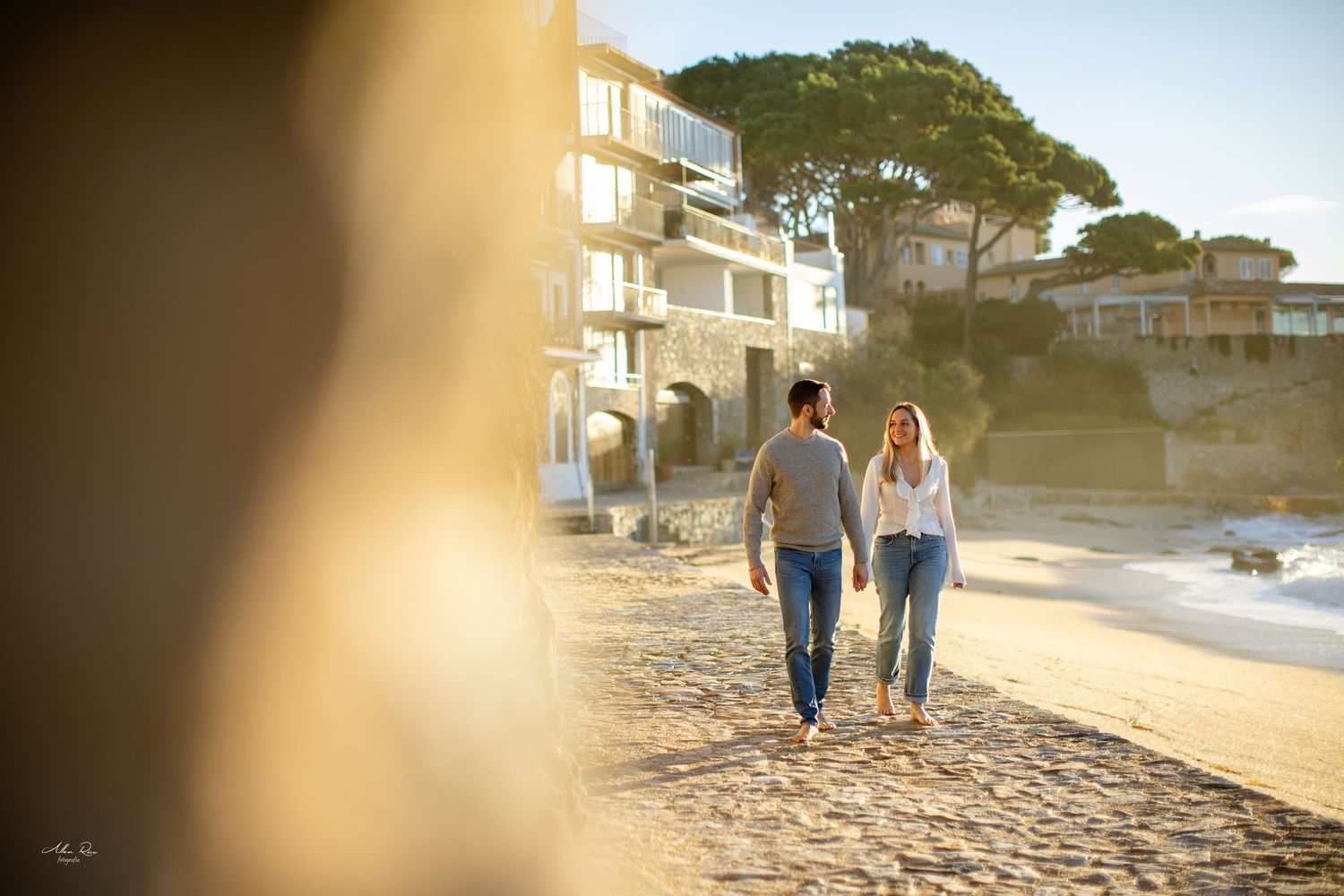 Couple walking hand in hand along a sunny beach with coastal buildings behind.