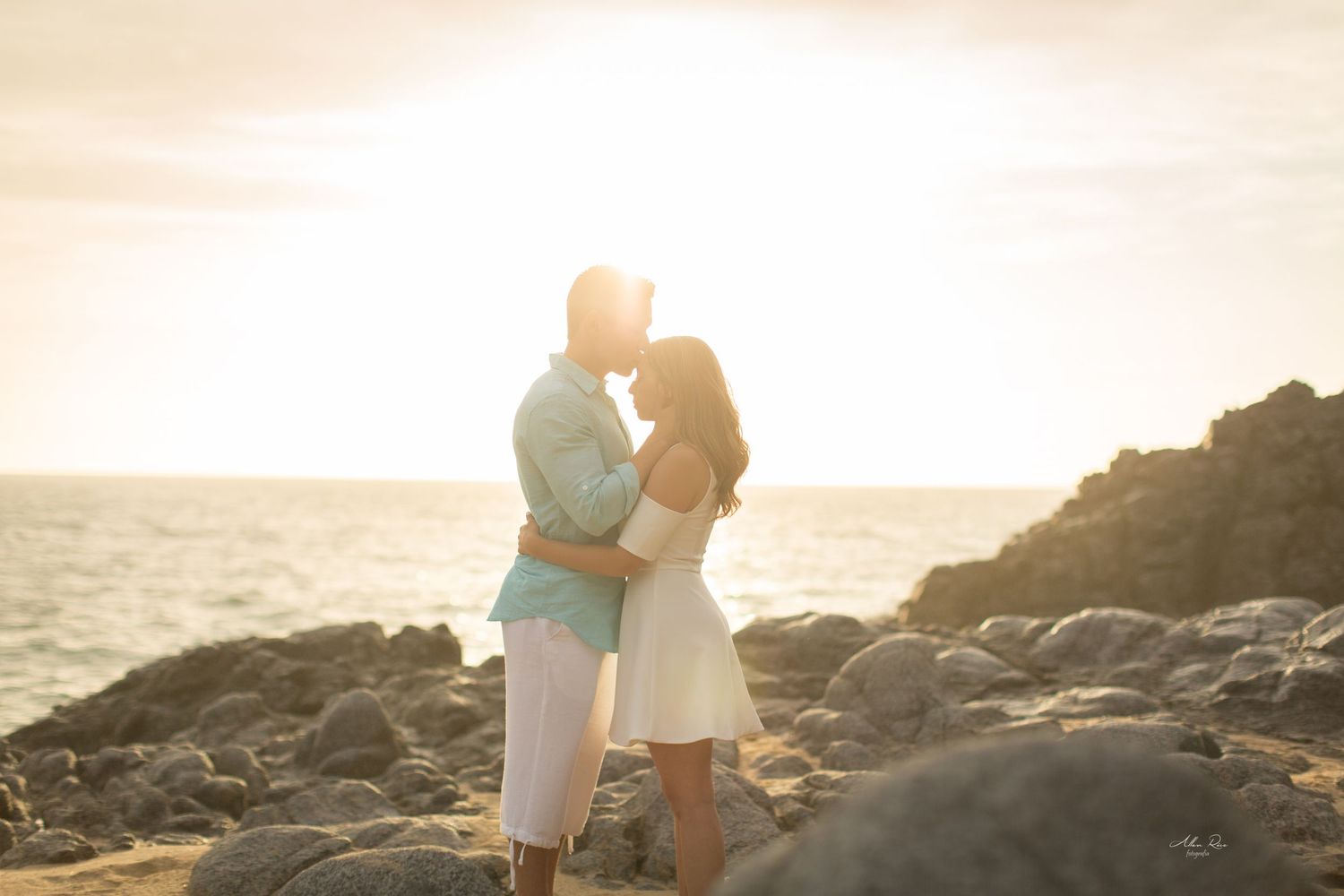Couple shares a sunset kiss on rocky beach with golden ocean light behind them.