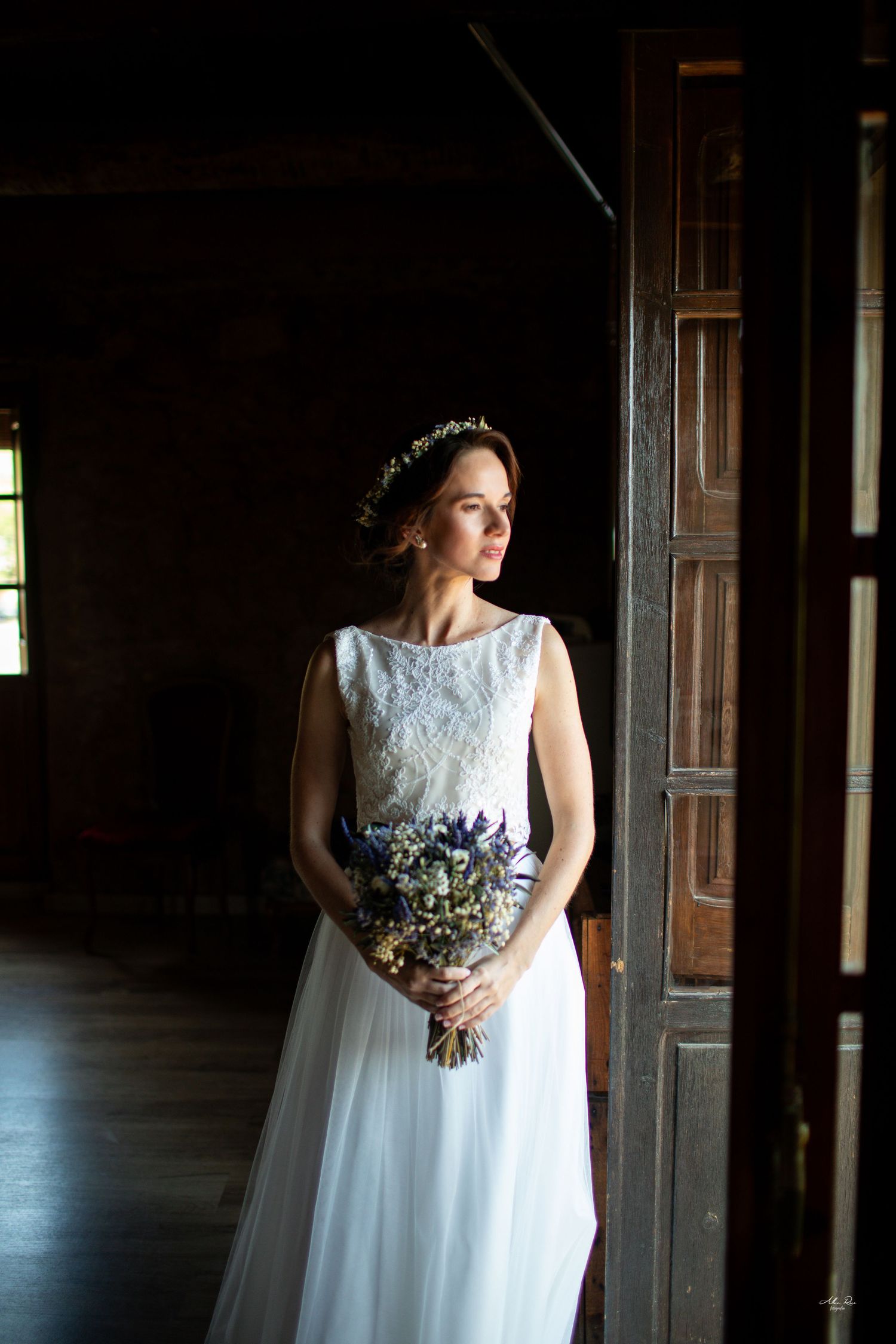 Bride in white lace gown holding wildflower bouquet stands by wooden doors in dark rustic interior.