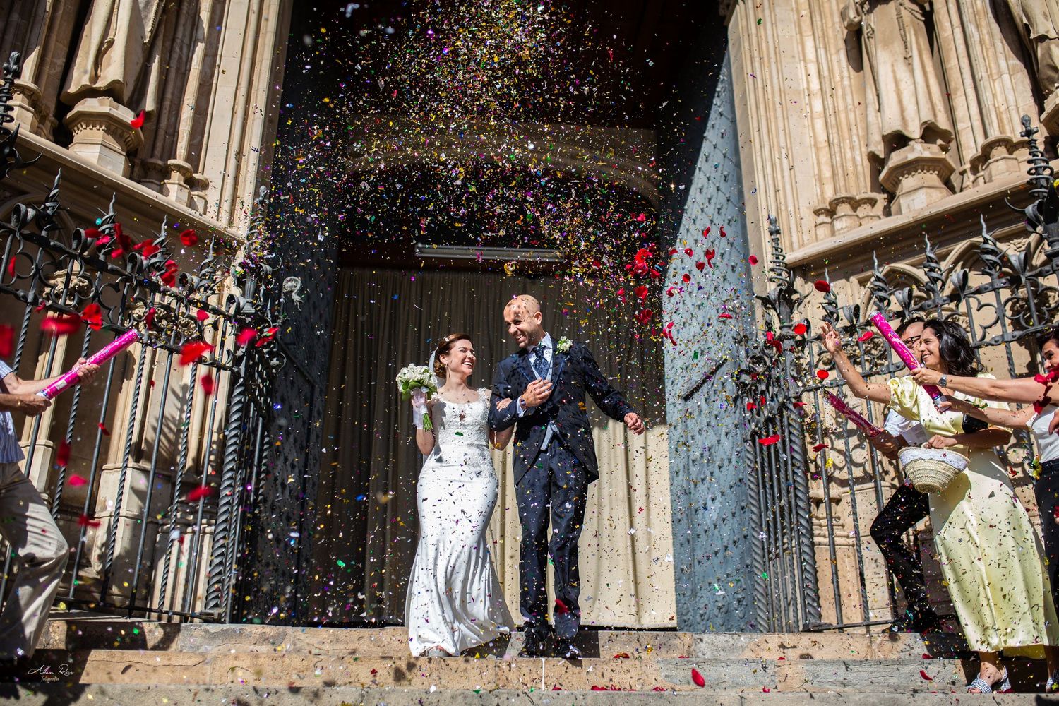 Joyful newlyweds exit church doors surrounded by colorful confetti celebration.