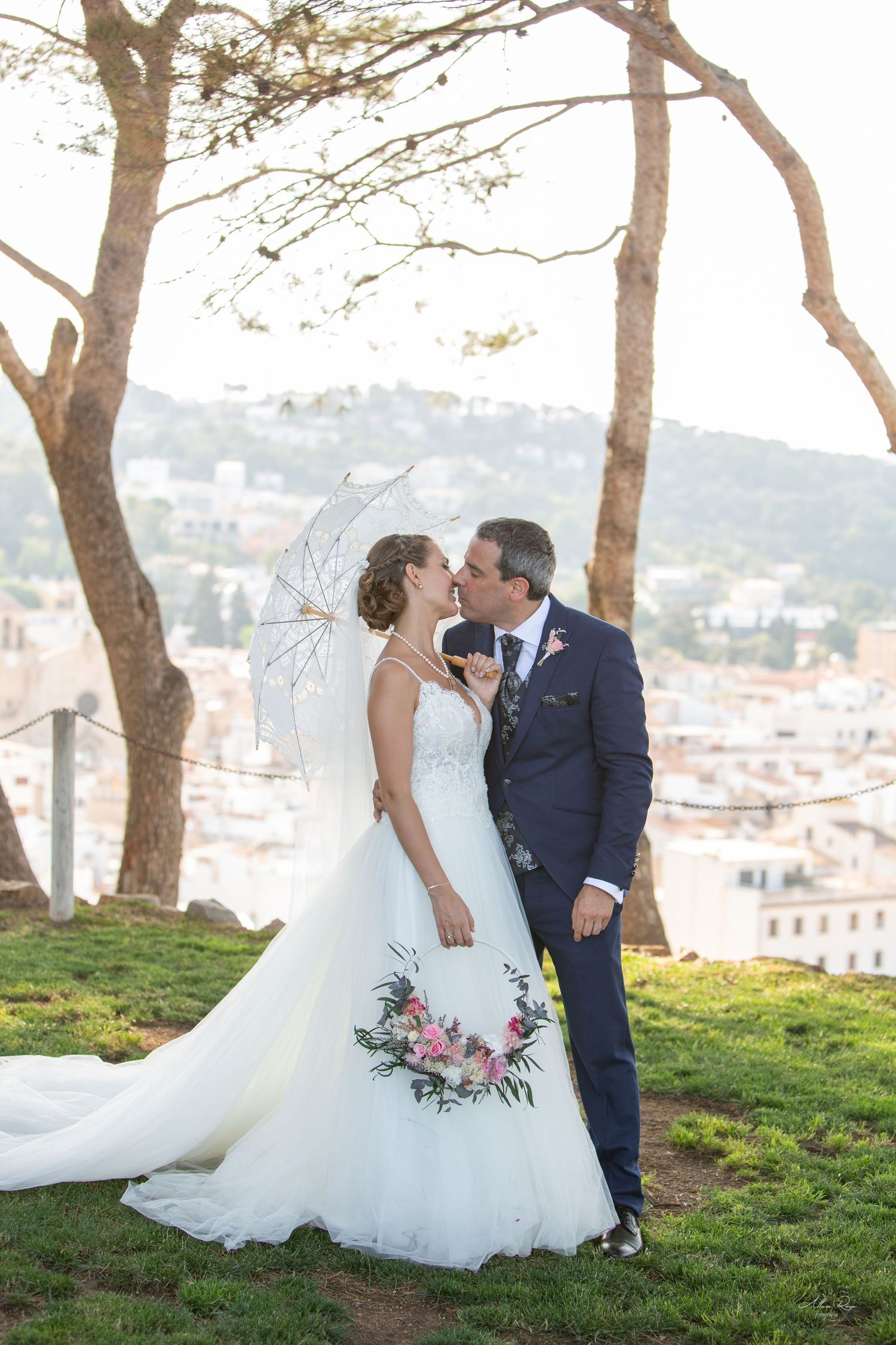 Bride and groom share a kiss on a hilltop garden with a picturesque town backdrop and lace parasol.