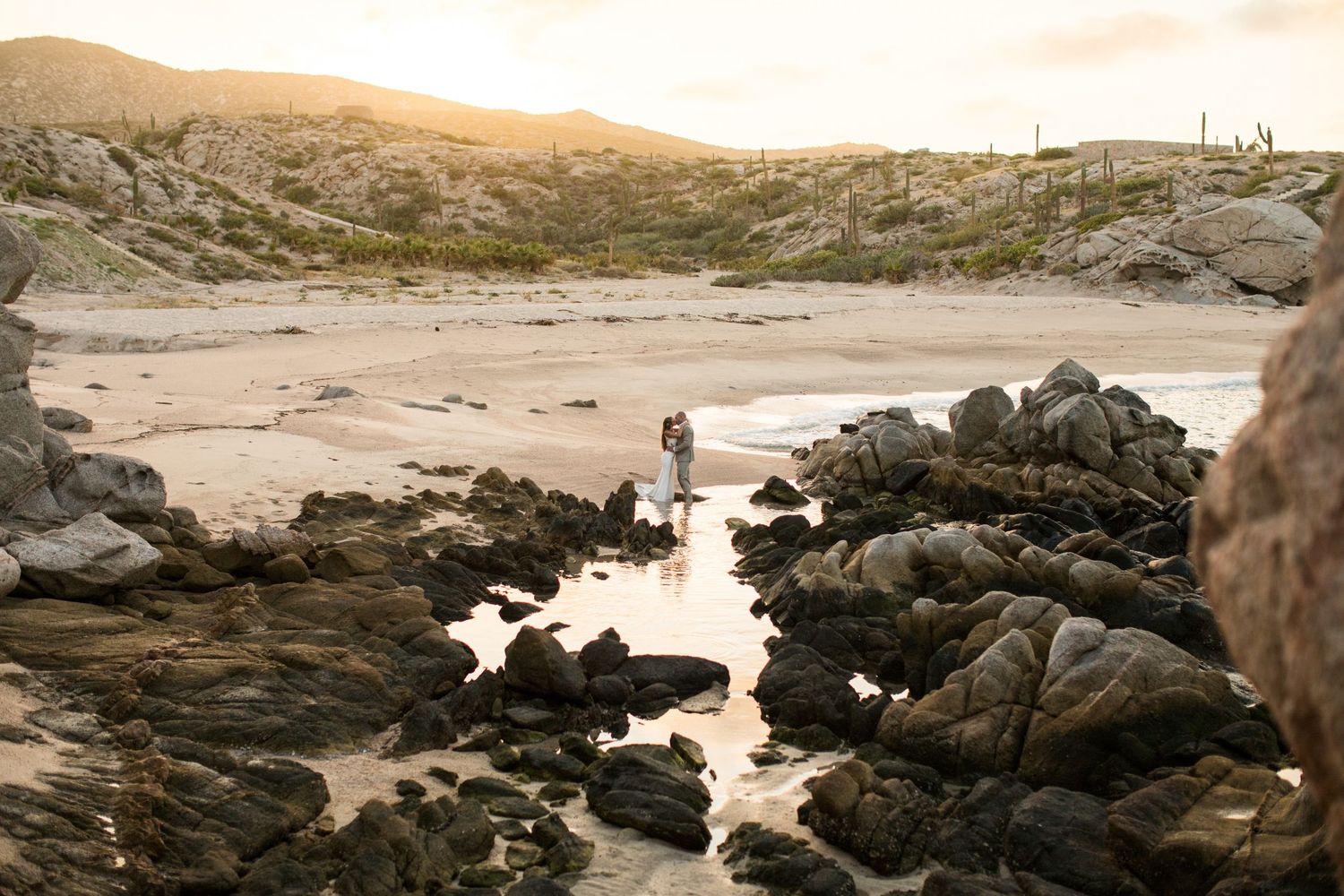 Couple embracing on a rocky beach at golden hour with desert hills in the background.
