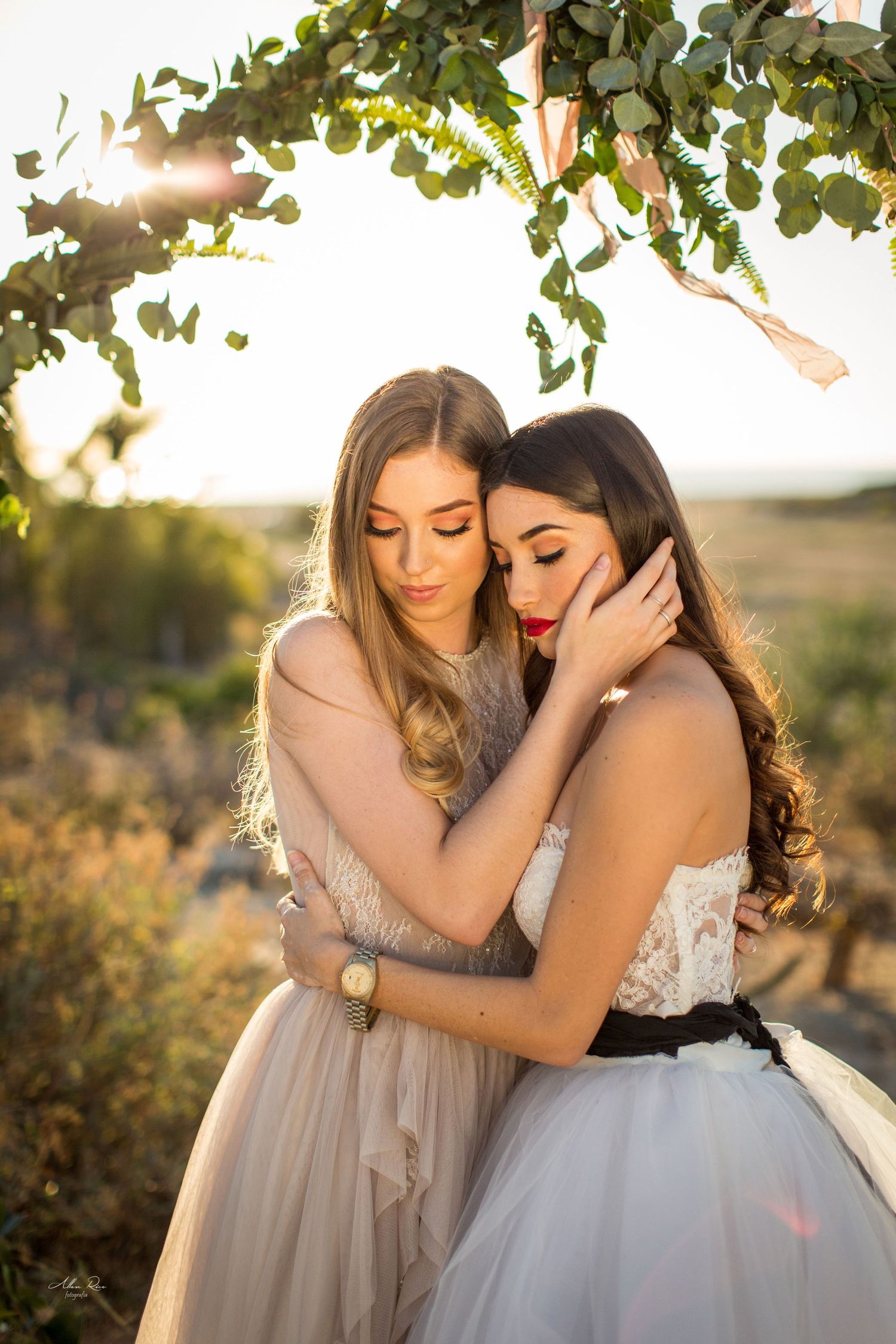 Two brides share an intimate embrace under a leafy wedding arch with warm sunset light in the background.