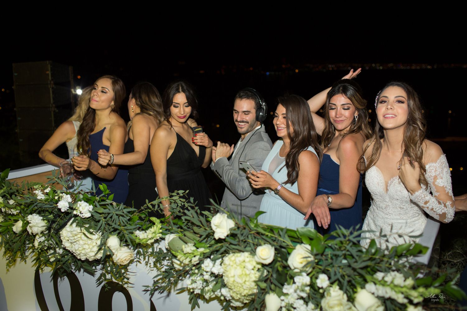 Wedding party poses with bride in white gown beside floral arrangements at night event.