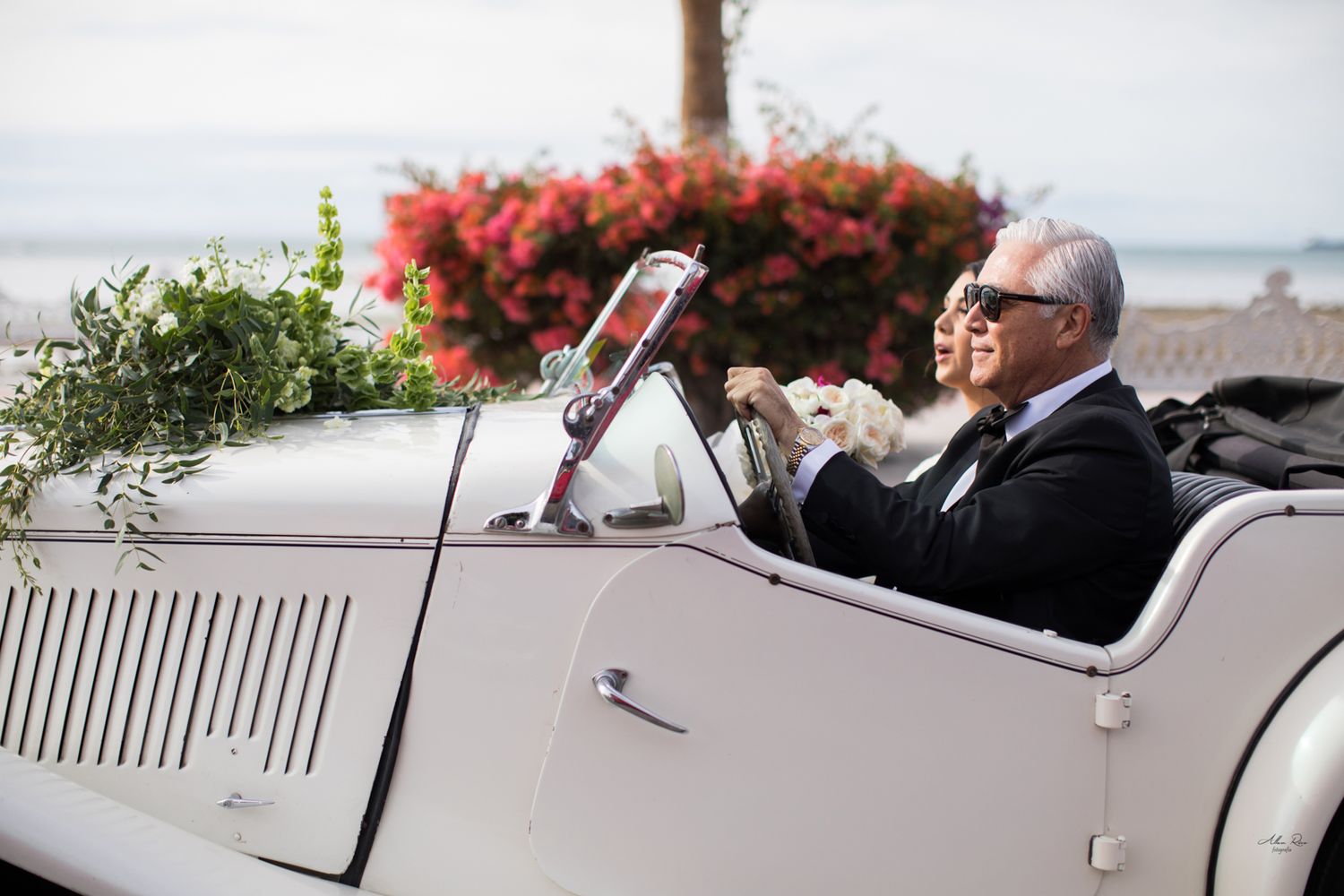 Couple riding in decorated vintage white convertible car with red floral backdrop outdoors.