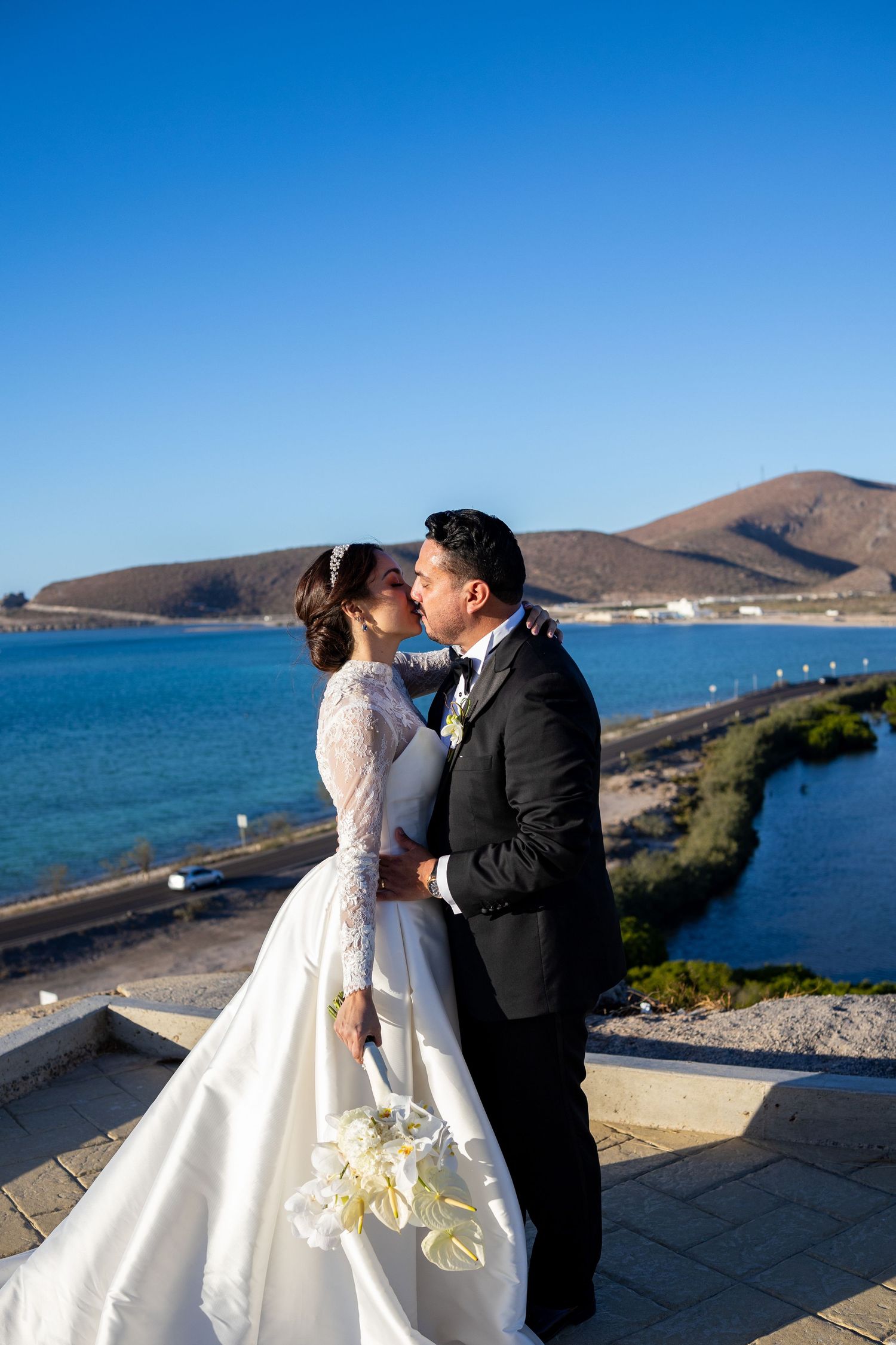 Bride and groom kissing with scenic blue bay and mountain backdrop on wedding day.