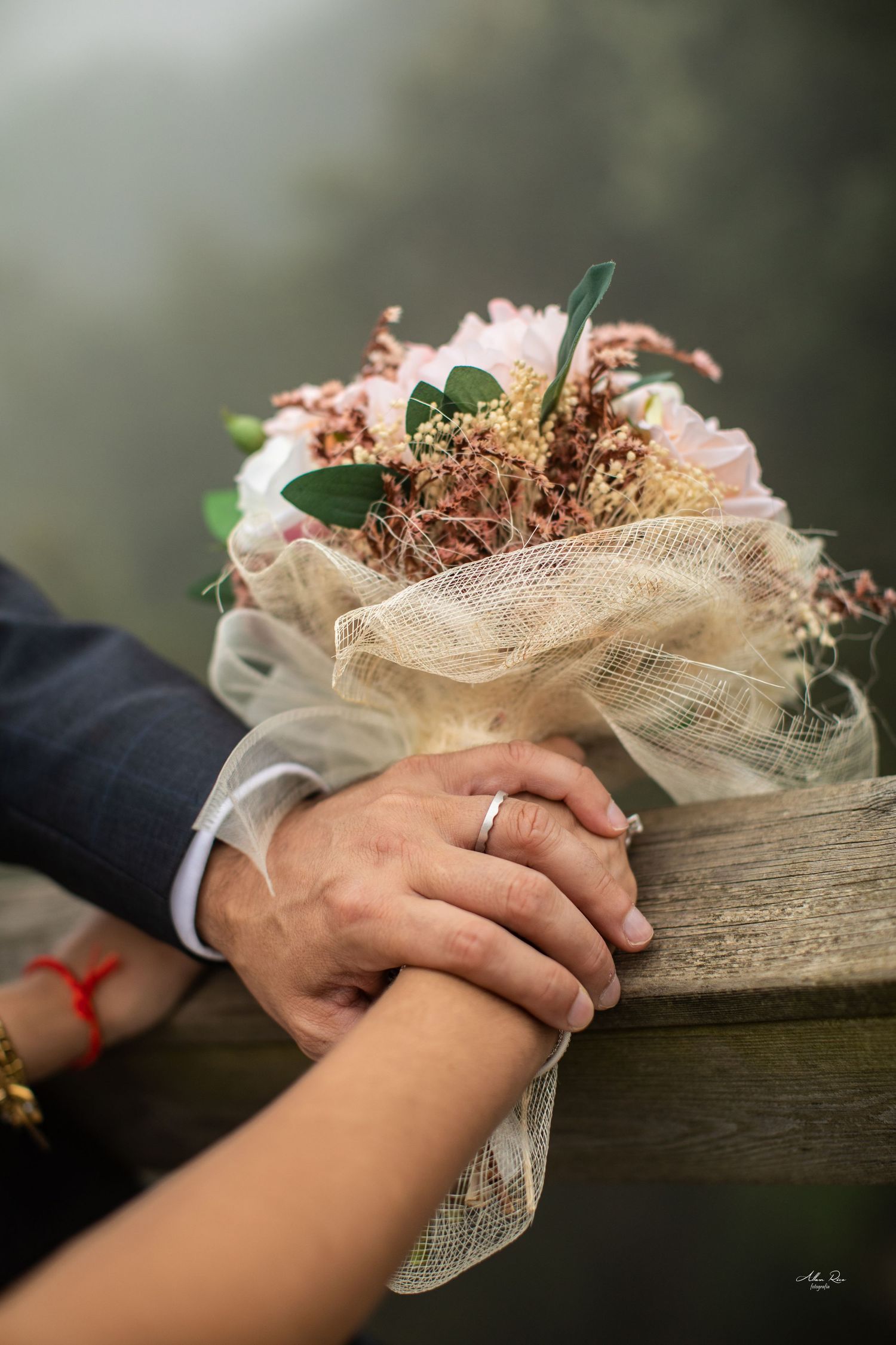 Close-up of newlyweds holding hands beside rustic bouquet of pink flowers.
