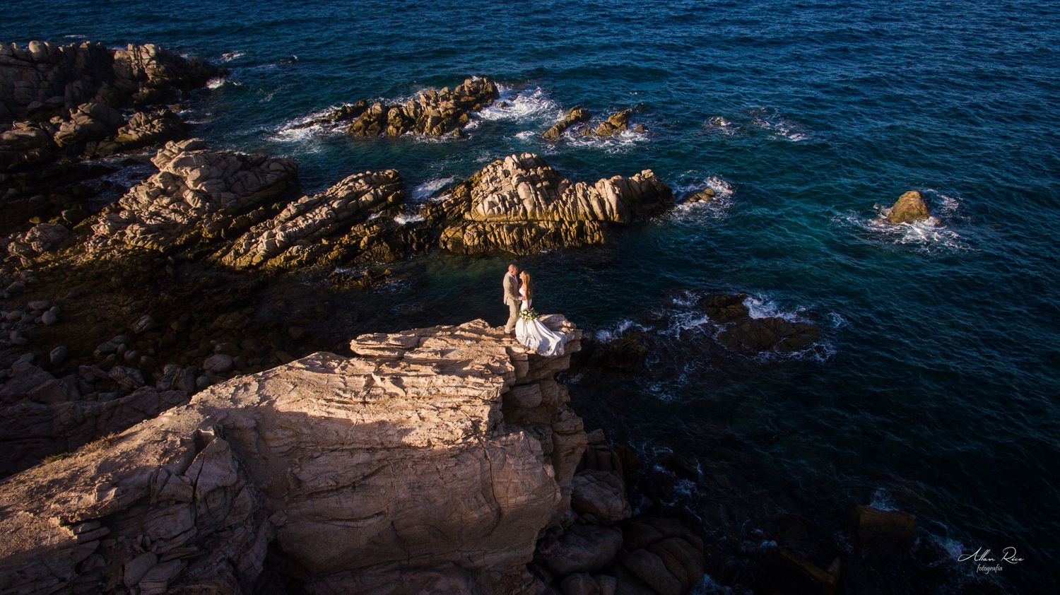 Bride in white gown stands alone on rocky coastal cliffs with turquoise ocean waves below.