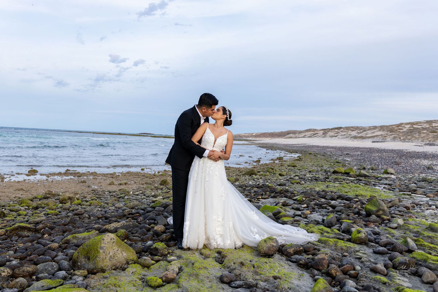 Couple shares romantic kiss on rocky shoreline with green moss, bride in flowing white gown with train.