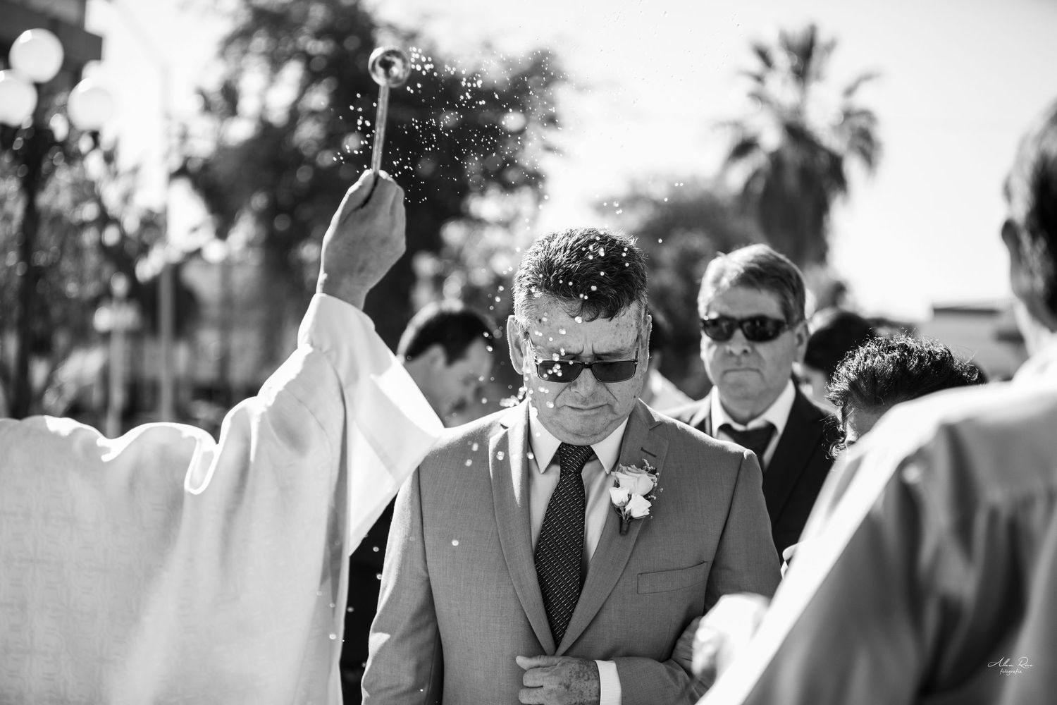 Black and white photo of priest blessing groom with holy water during outdoor wedding ceremony.