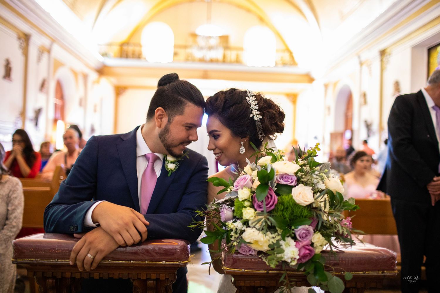 Bride and groom share an intimate moment at the altar, with a beautiful purple and white bouquet in a church.