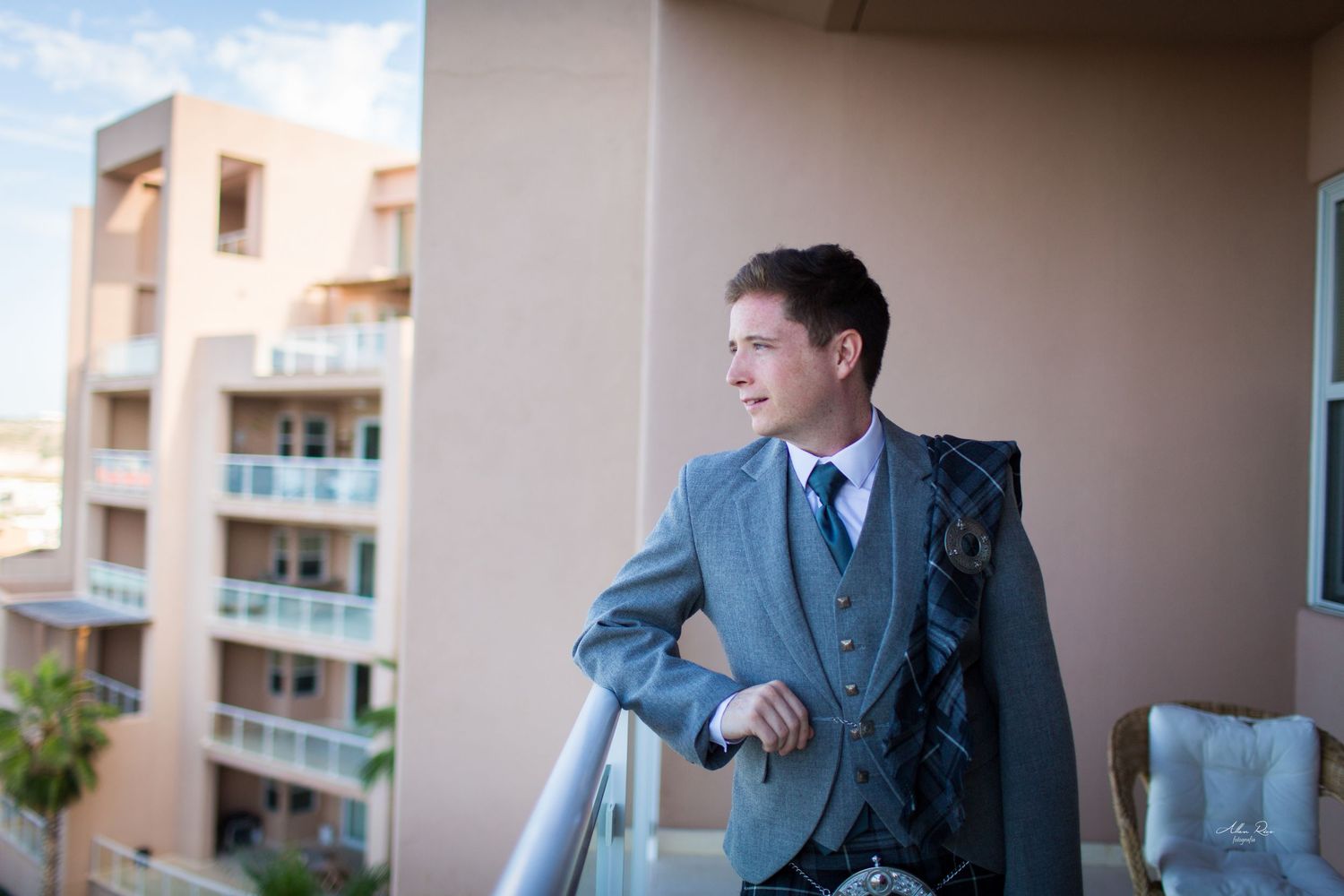 Dapper groom in grey suit and kilt stands on hotel balcony gazing at city views.