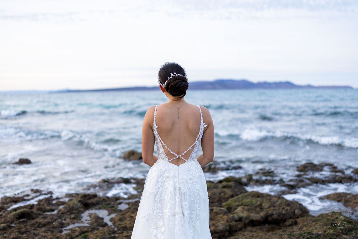 Bride in backless lace wedding gown standing before the ocean on rocky shore.