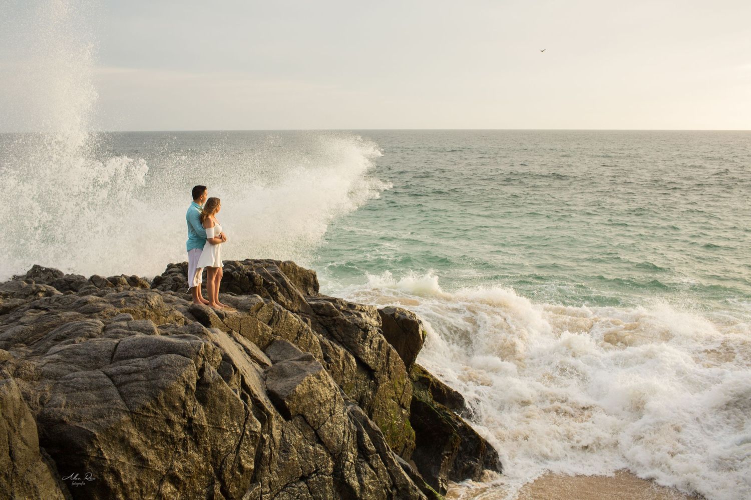 Couple stands on coastal rocks as powerful ocean waves crash dramatically behind them.