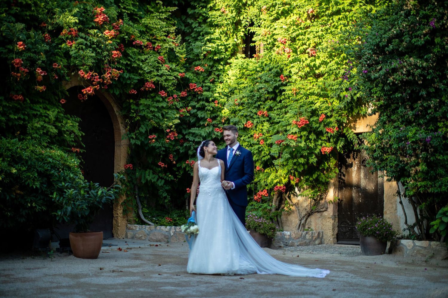 Elegant bride and groom posing in a lush courtyard with vibrant red climbing flowers.