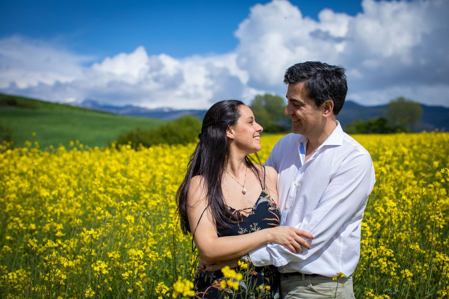 Couple holding hands and smiling in vibrant yellow flower field with mountains and blue sky.