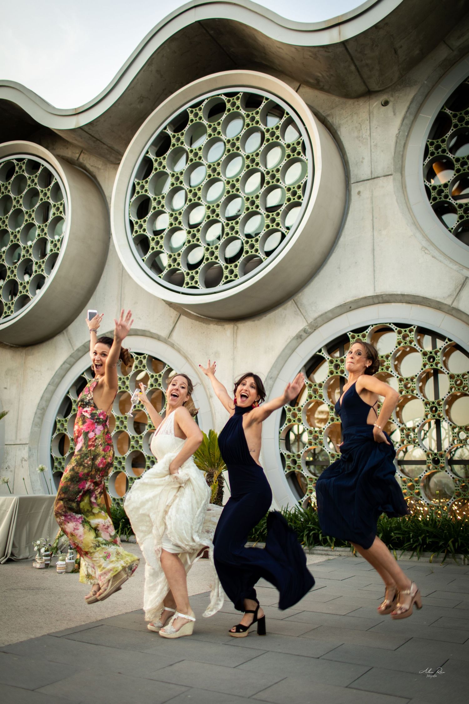 Bride and bridesmaids joyfully jumping outside modern building with large circular decorative windows.