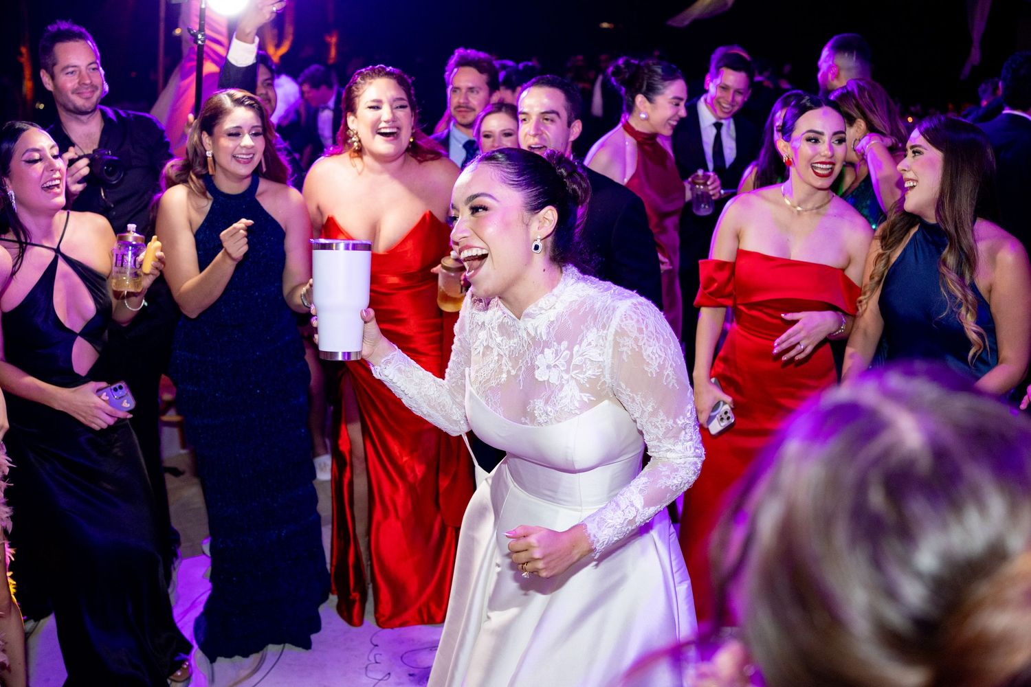 Radiant bride in long-sleeve white gown laughing and celebrating with guests at her wedding reception.