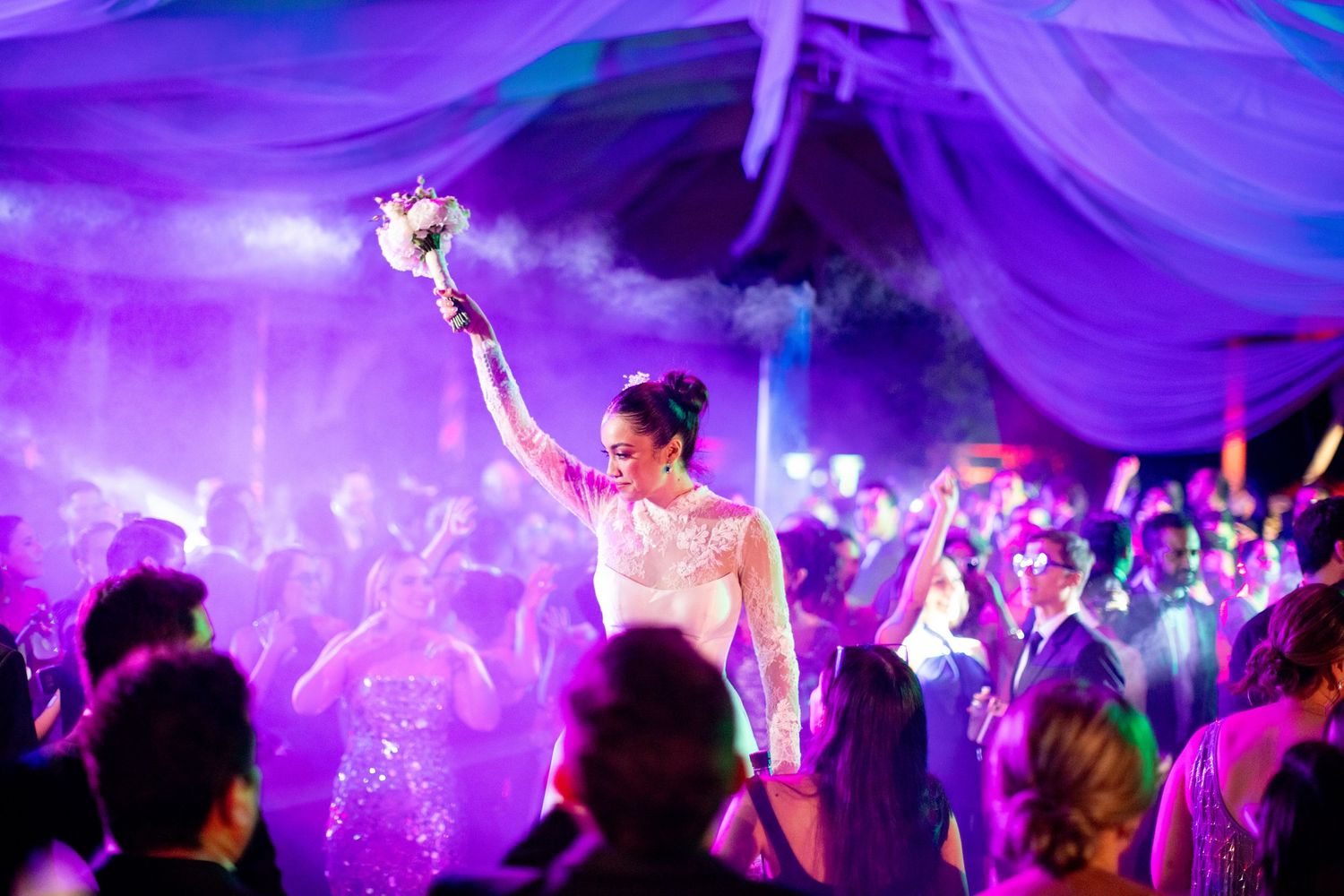Bride triumphantly raises bouquet above crowd on a smoky, purple-lit wedding reception dance floor.