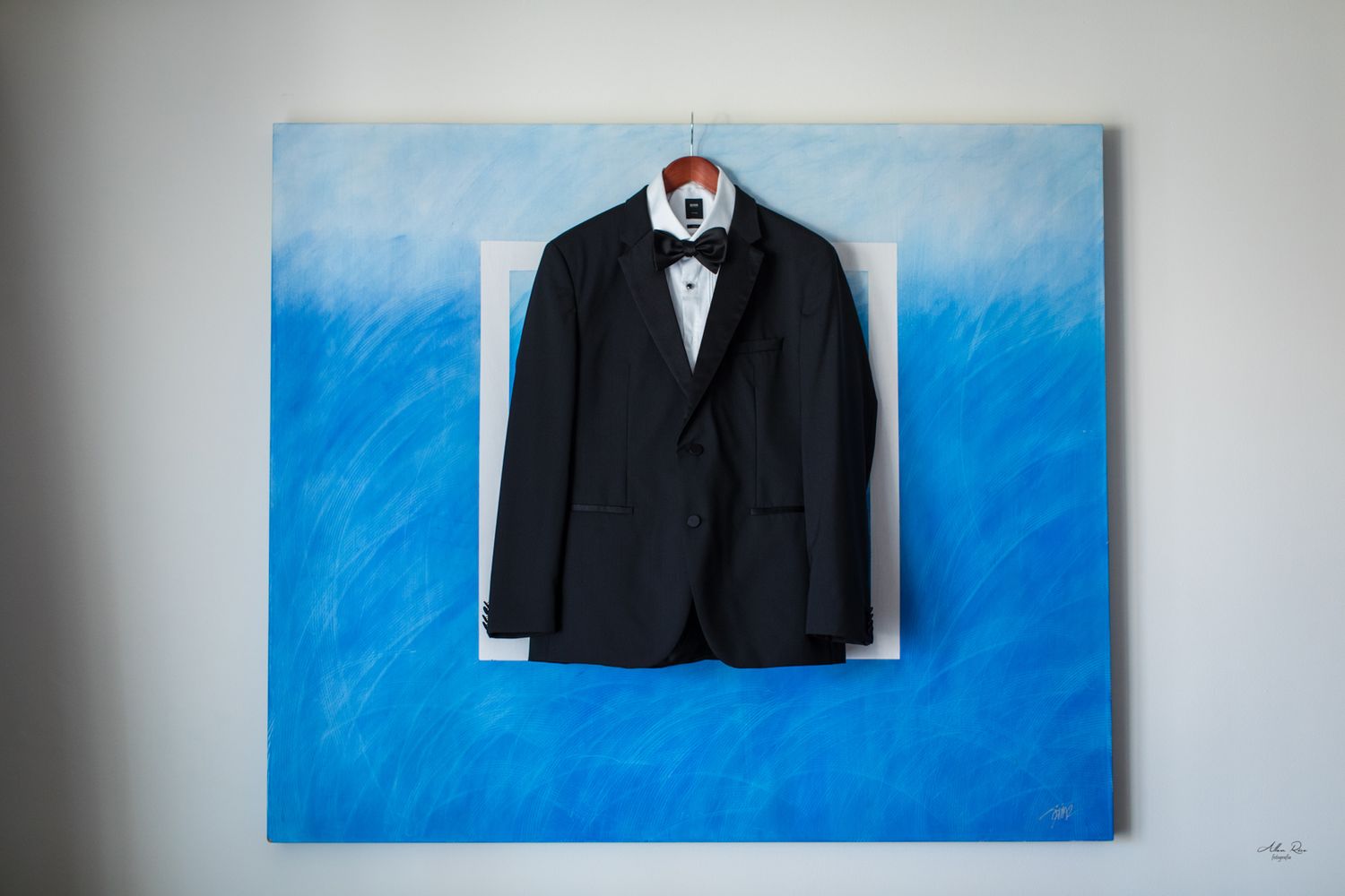 Black tuxedo jacket with bow tie hanging on a blue painted canvas wall.