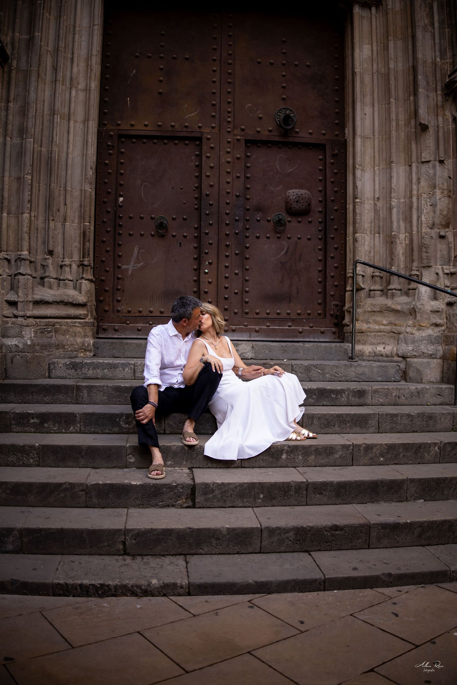 A couple in white attire sits intimately on stone steps before a grand historic wooden door in Barcelona.