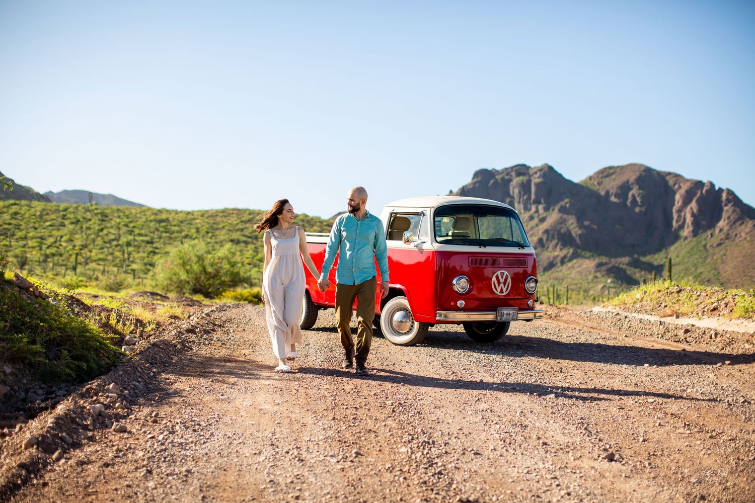 Couple walking hand in hand on a desert road by a red VW van.