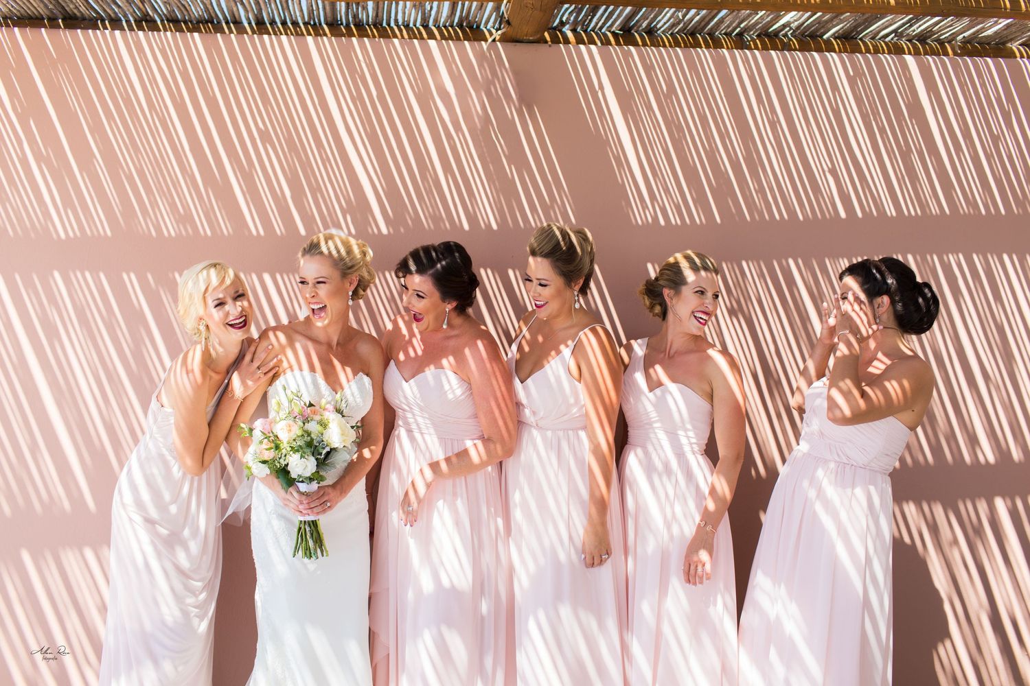 Bride and four bridesmaids in blush gowns laughing together under striped light shadows.