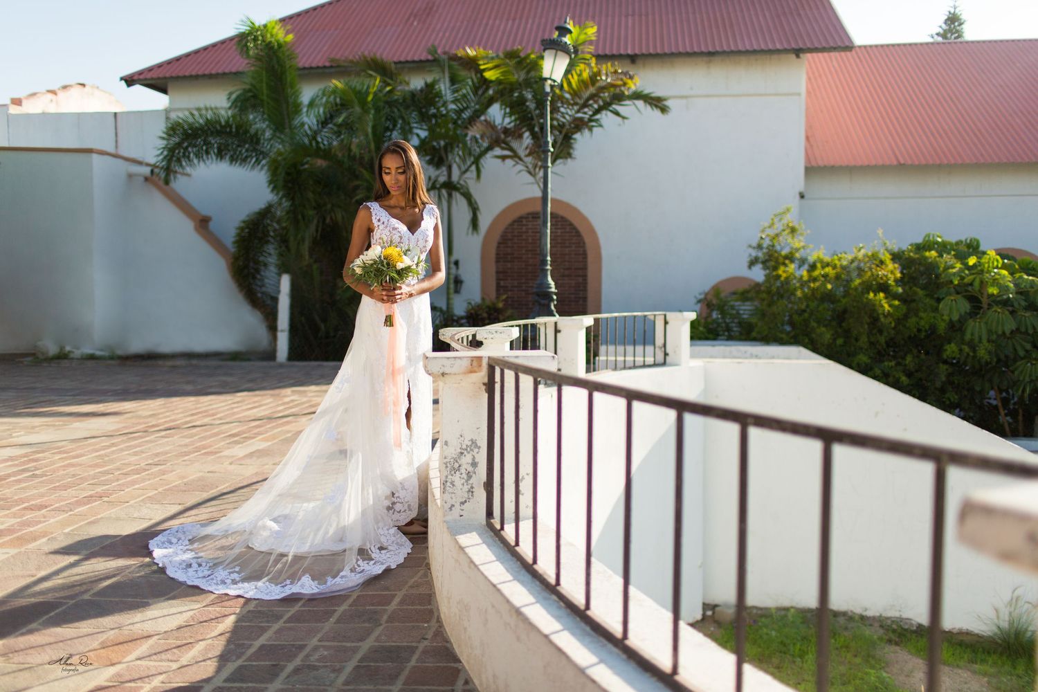 Bride in white lace wedding gown with bouquet stands on sunny terrace outside colonial building.