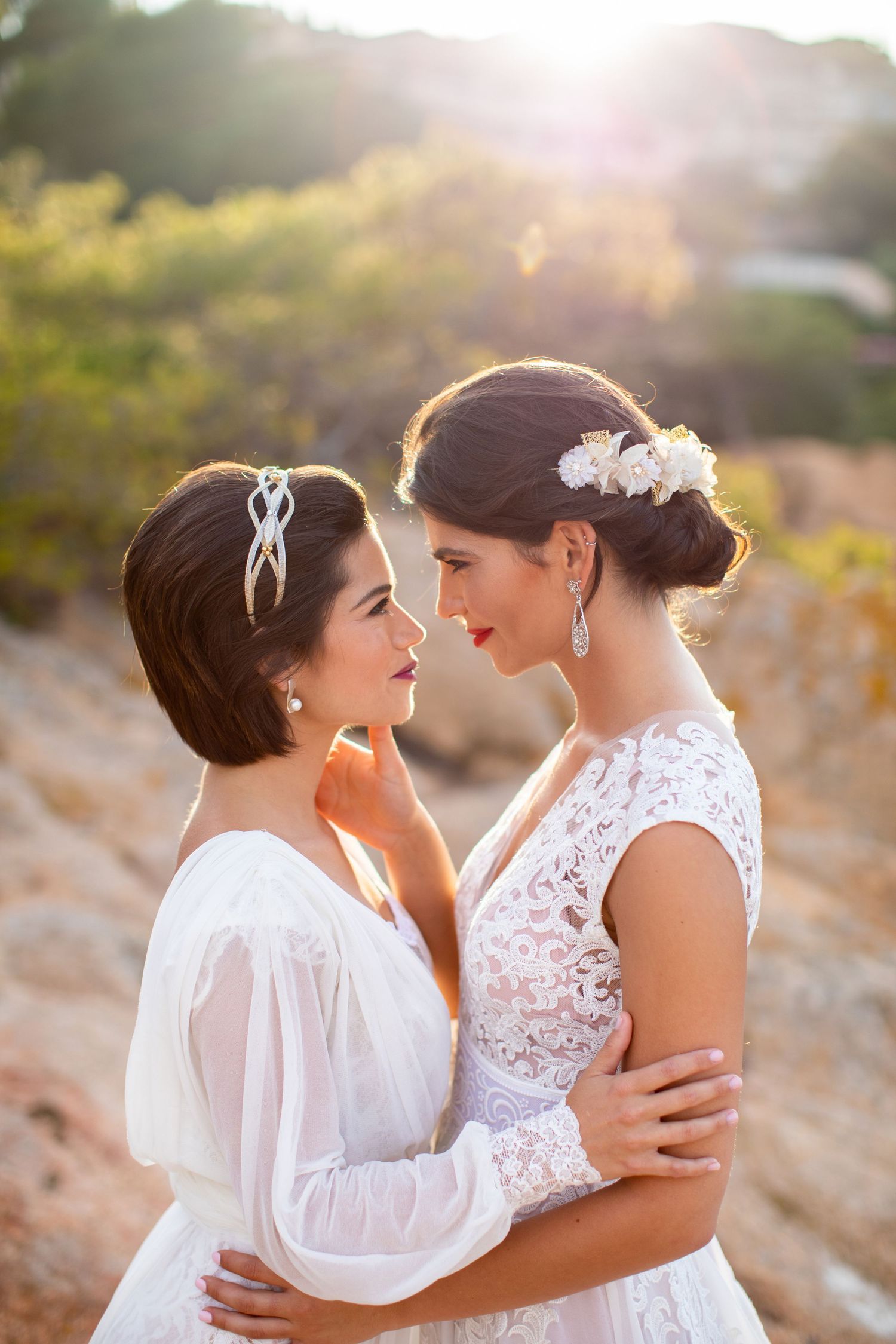 Two brides in elegant white wedding gowns embrace romantically at sunset on rocky terrain.