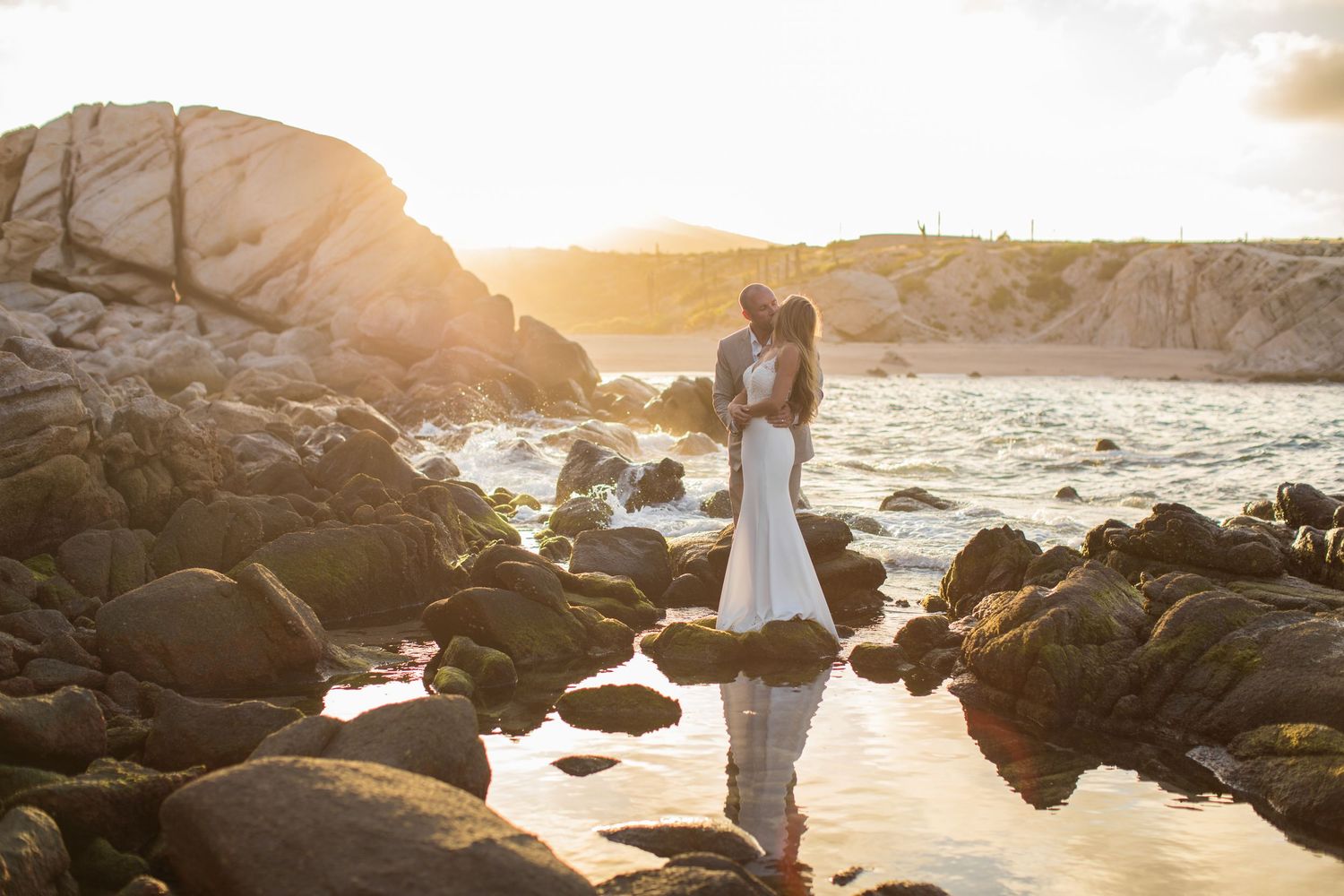 Couple shares a passionate kiss on rocky shoreline bathed in warm golden sunset light.