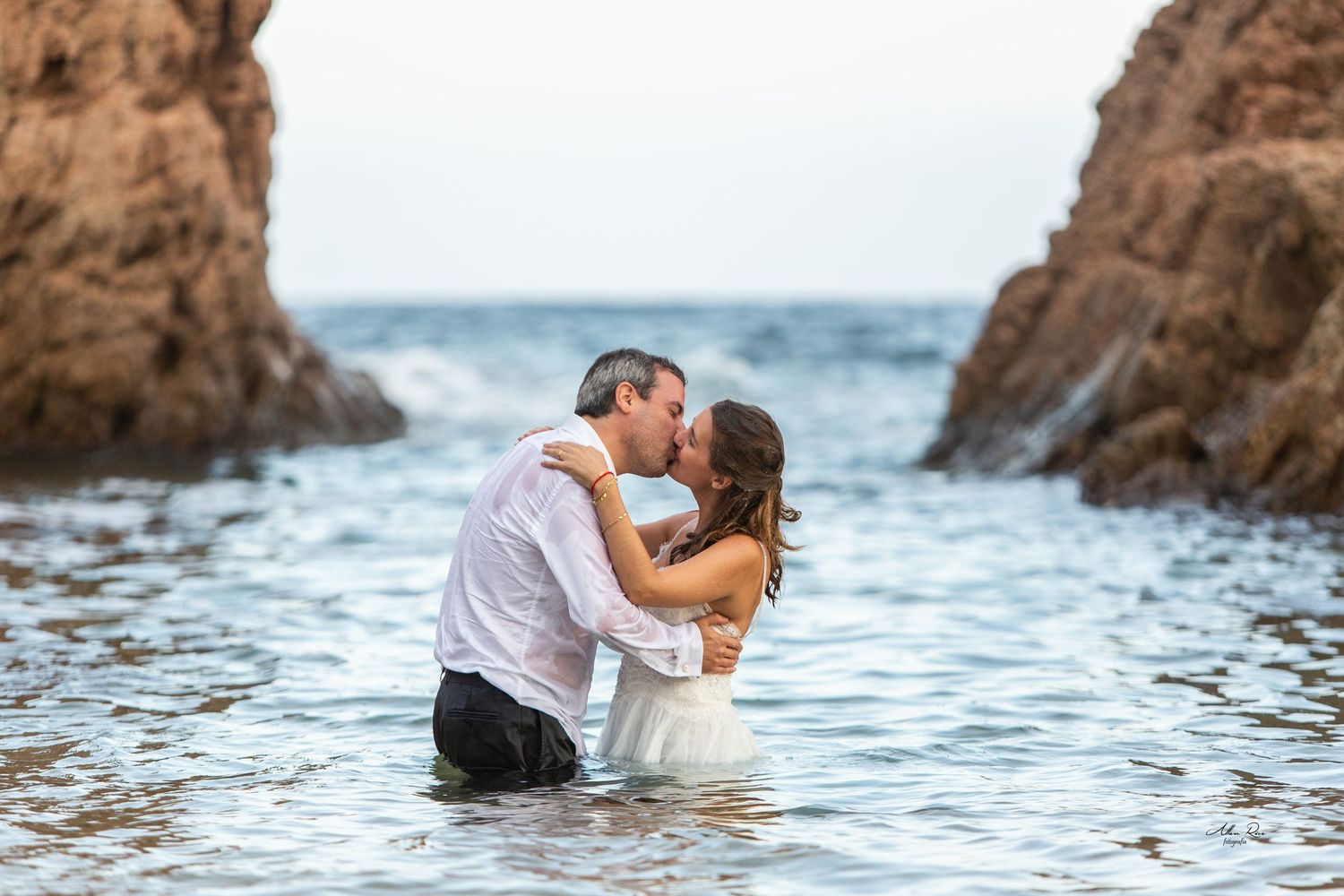 Couple shares a passionate kiss standing in ocean water between dramatic rock formations.