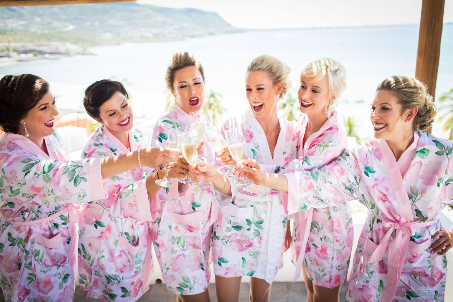 Laughing bridesmaids in pink floral robes enthusiastically clinking champagne glasses with scenic backdrop.