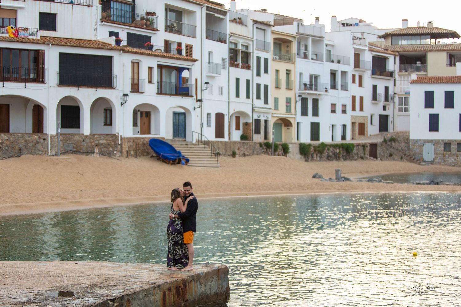Couple embracing on a waterfront pier with colorful Mediterranean-style buildings lining the beach shore.
