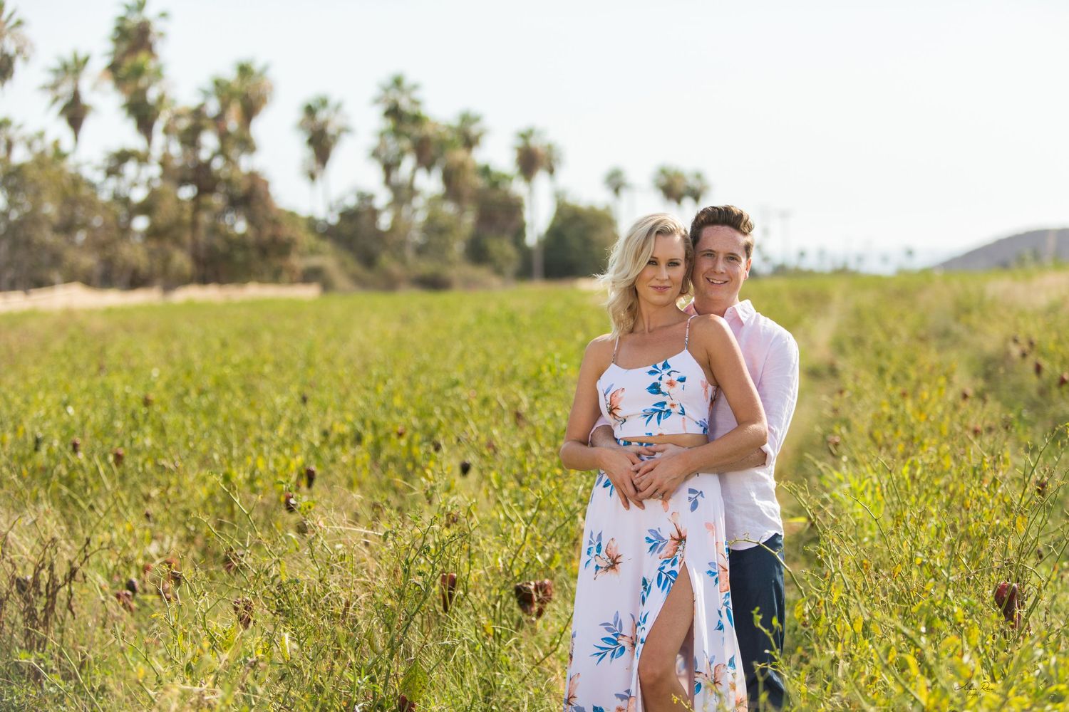 Couple embracing in sunny green meadow with palm trees, woman in floral sundress, man in white shirt.