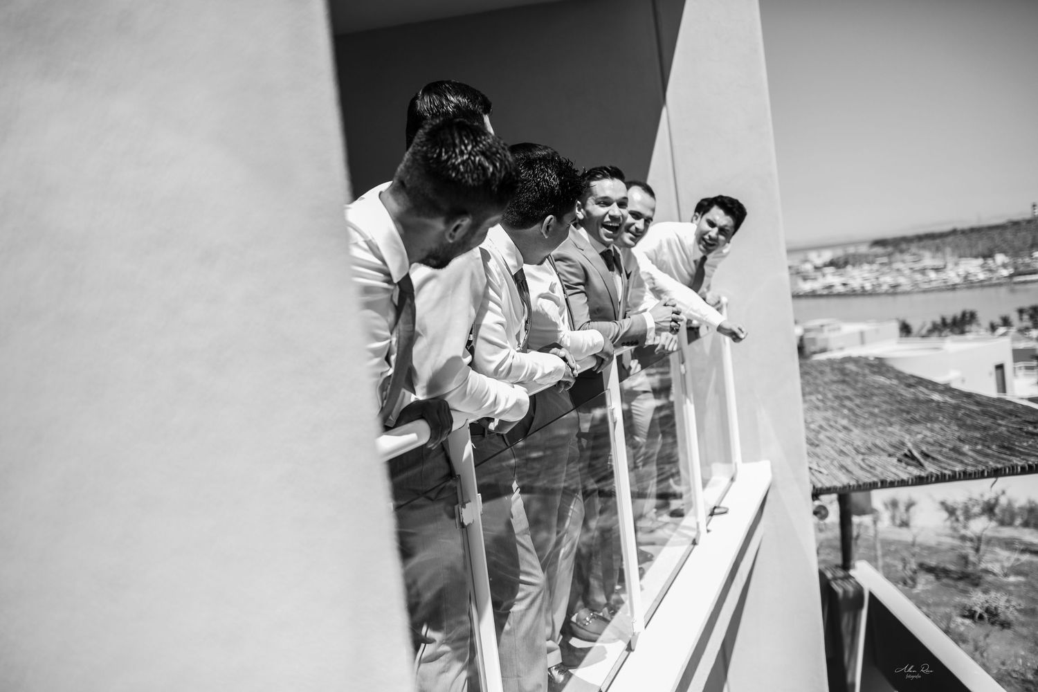 Groomsmen laughing and leaning on a balcony railing with a scenic town view in a black and white wedding photo.