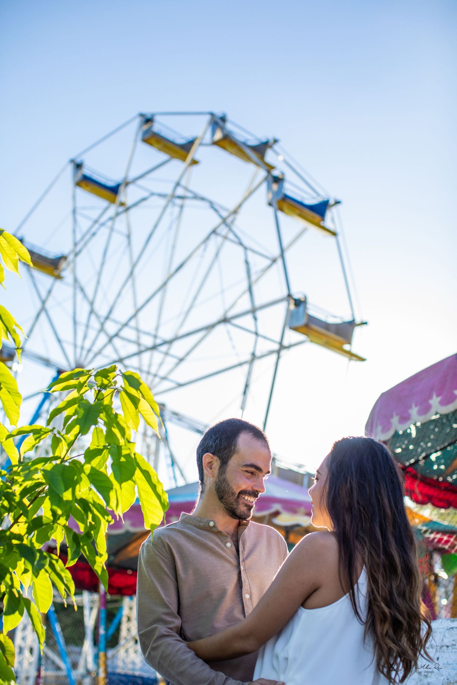 Happy couple embracing and laughing at a colorful amusement park with a ferris wheel in the background.