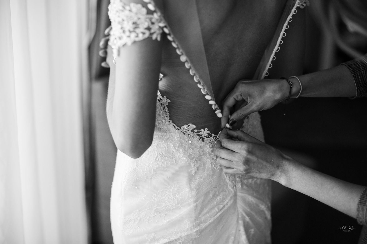 Black and white close-up of hands fastening buttons on a lace backless wedding dress.