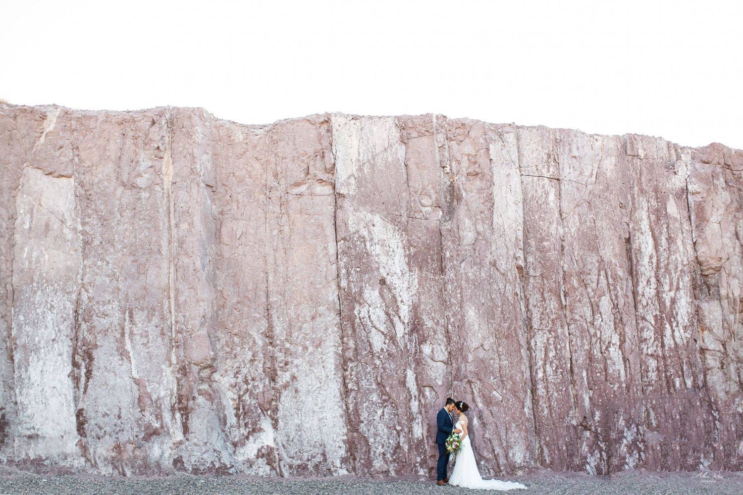 Bride and groom share a kiss against a dramatic red rock cliff face in a minimalist outdoor setting.