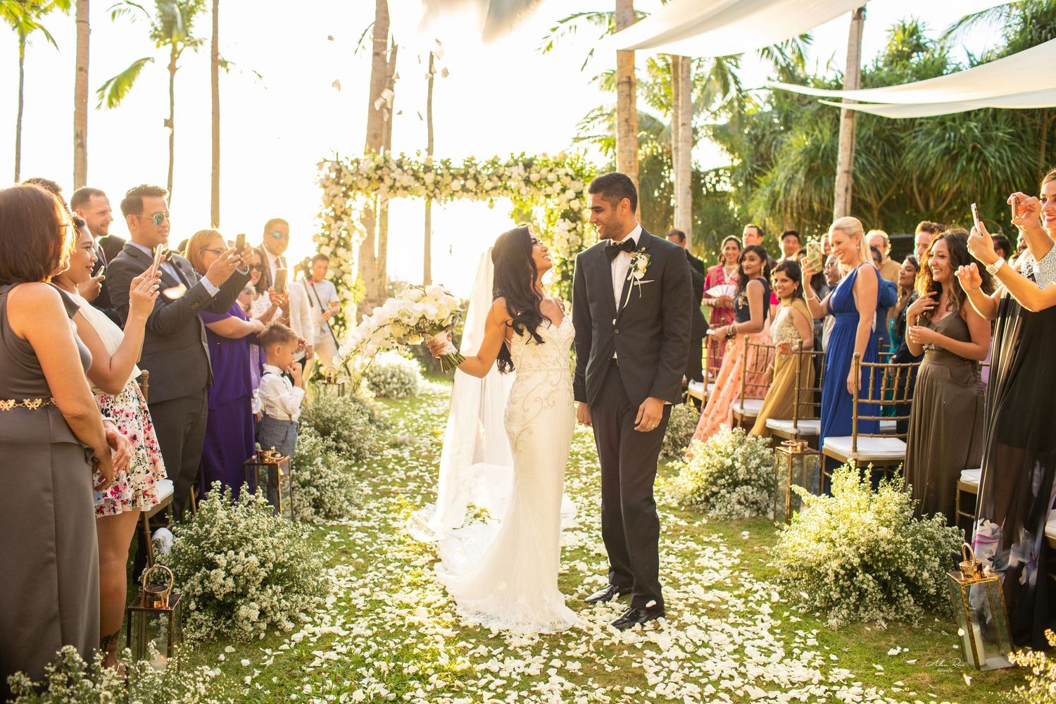 Bride and groom share first dance at outdoor wedding ceremony with floral arch and petal-lined aisle.