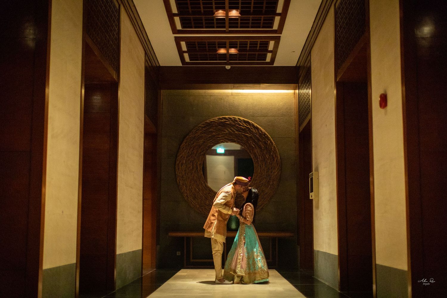Indian couple sharing a romantic kiss in an elegantly lit hotel corridor at night.