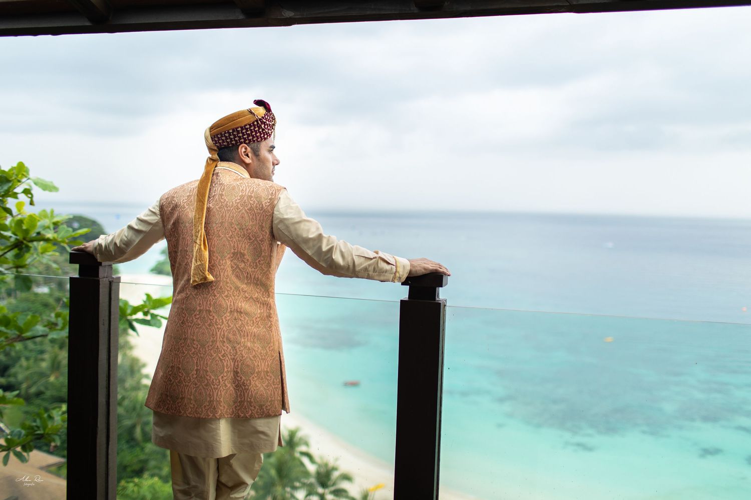 Indian groom in traditional attire overlooking turquoise ocean from a tropical resort balcony.
