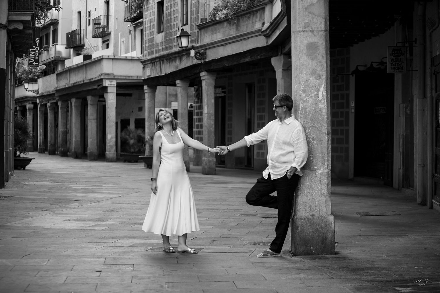 Black and white photo of couple holding hands playfully beside a stone column in a plaza.