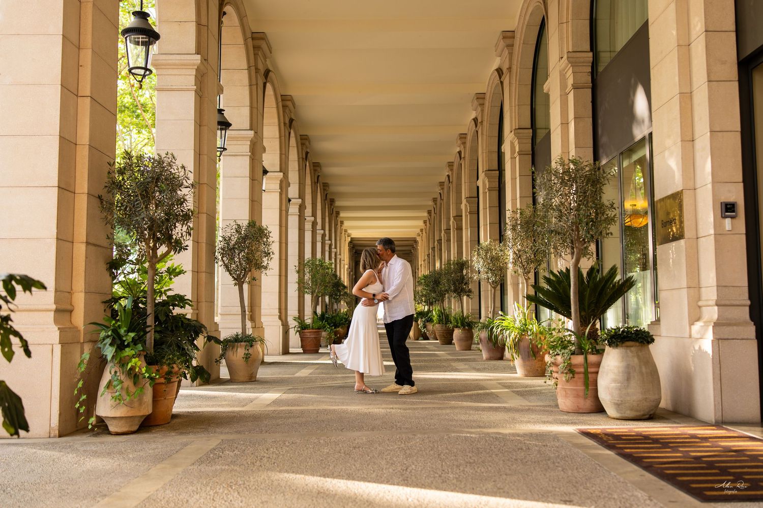 Couple dancing romantically in a sunlit arcade corridor lined with potted olive trees.