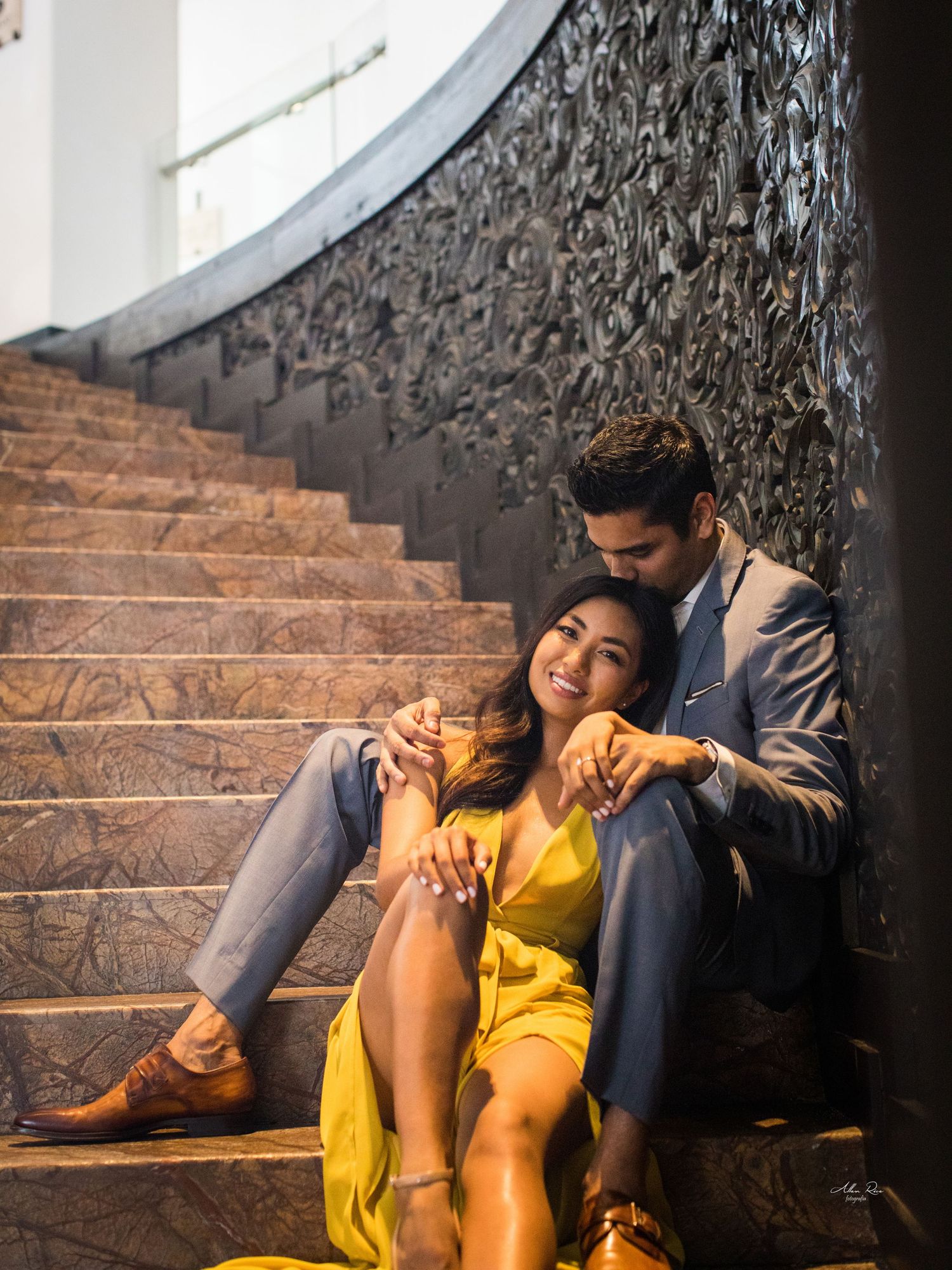 Couple sitting together on ornate marble staircase, woman in yellow dress leaning against partner.