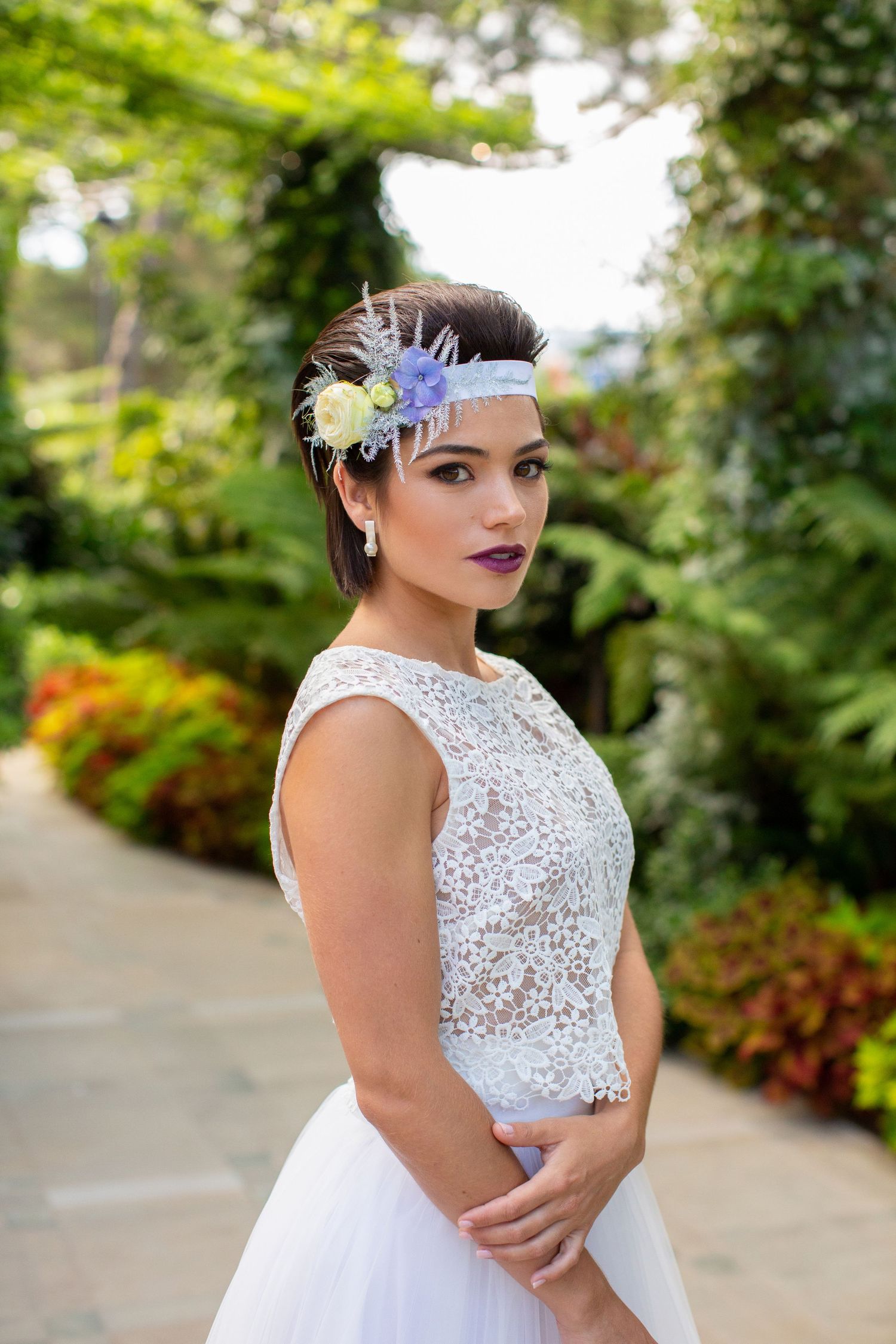 Bride wearing a lace cap-sleeve wedding dress and floral headpiece poses in a lush green garden setting.