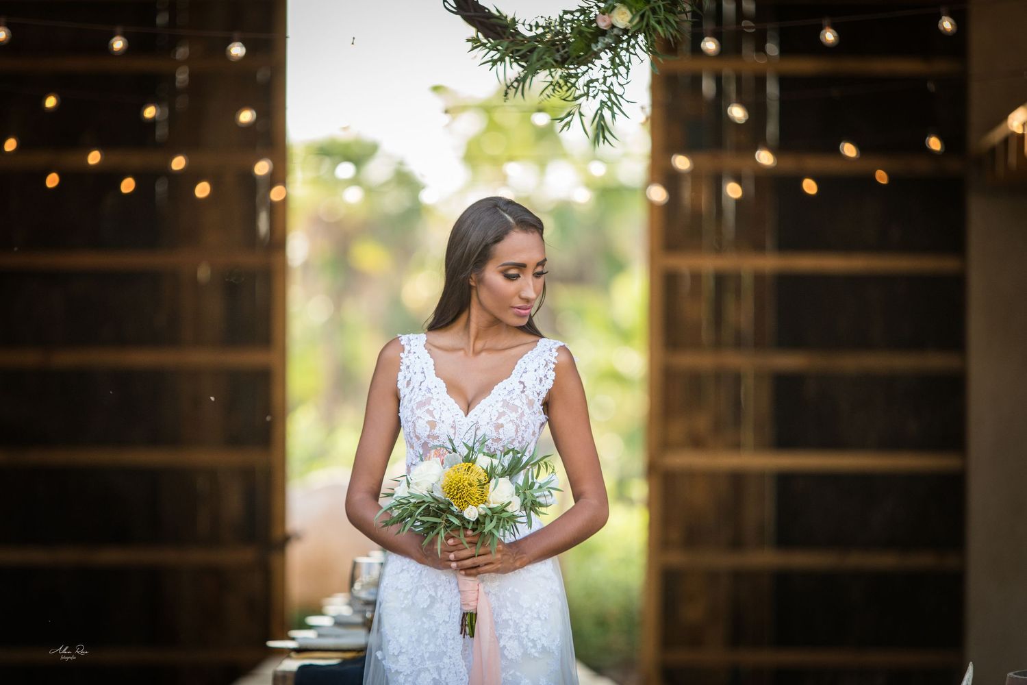 Bride in lace wedding gown holding yellow and white bouquet standing in barn doorway with string lights.
